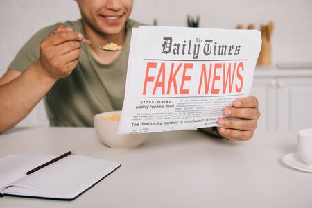 cropped view of smiling man eating flakes while reading fake news newspaper in kitchenの写真素材