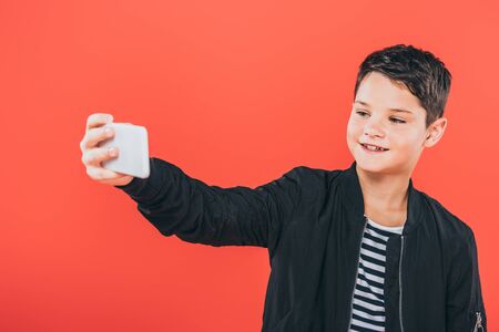 smiling kid in jacket taking selfie isolated on redの写真素材