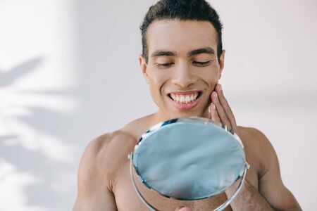 handsome mixed race man touching face with hand while smiling and looking at mirrorの写真素材