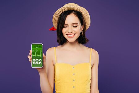 smiling girl in straw hat holding smartphone with healthcare app isolated on purple