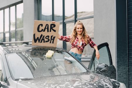 positive young woman holding carton board with car wash lettering and standing near automobileの写真素材