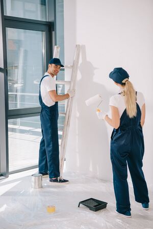 handsome painter standing near ladder while pretty colleague holding paint rollerの写真素材