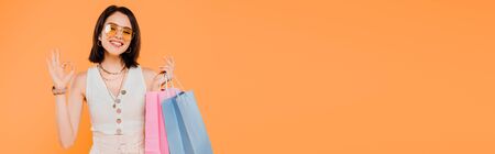 happy fashionable girl in sunglasses holding shopping bags and showing okay sign isolated on orangeの写真素材