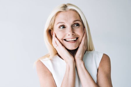 happy blonde mature woman with hands near face looking away isolated on grey