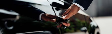 cropped view of african american businessman in suit opening car door at sunny day, panoramic shot