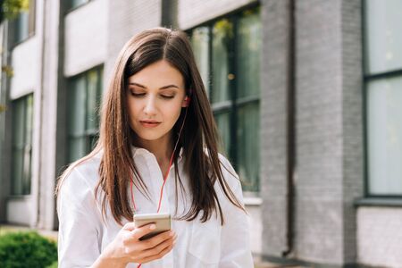 attractive young woman in white shirt holding smartphone and listening music in earphones on streetの写真素材