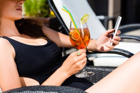 cropped view of girl in swimming suit with cocktail on sunbed using smartphoneの写真素材