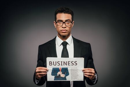 handsome african american businessman in glasses and suit holding business newspaper on dark backgroundの写真素材