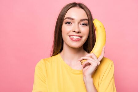 attractive smiling girl holding banana and looking at camera isolated on pinkの写真素材