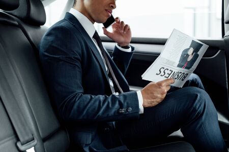 partial view of african american businessman reading business newspaper and talking on smartphone in carの写真素材