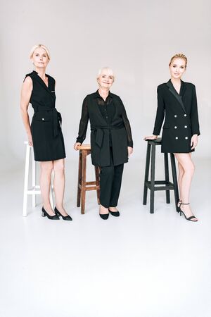 full length view of fashionable three-generation blonde women in total black outfits near chairs isolated on greyの写真素材