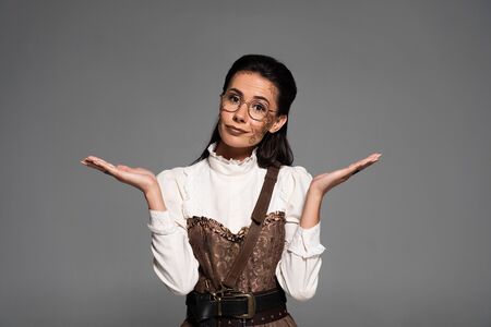 confused steampunk woman in glasses showing dubium gesture isolated on grey