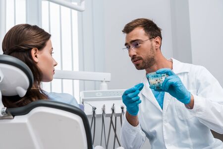 selective focus of attractive woman looking at handsome dentist in glasses holding tooth modelの写真素材