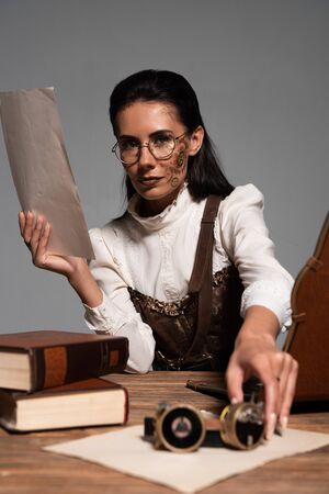 steampunk woman holding document and goggles at workplace isolated on grey