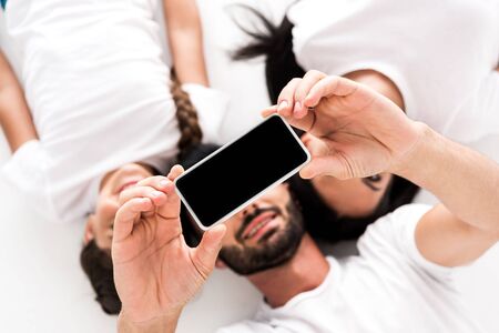 top view of happy bearded man holding smartphone with blank screen near wife and kid isolated on white の写真素材