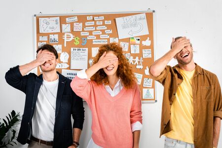 three smiling friends obscuring faces with hands in office