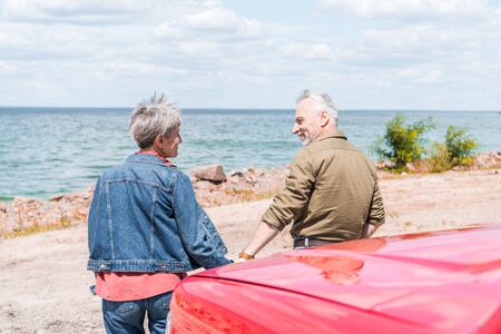 back view of senior couple standing near red car and looking at each other at beachの写真素材
