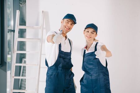 young painters in overalls showing thumbs up while smiling at cameraの写真素材