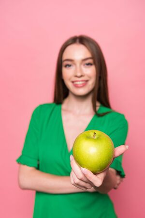 selective focus of attractive young woman holding green apple and smiling isolated on pinkの写真素材