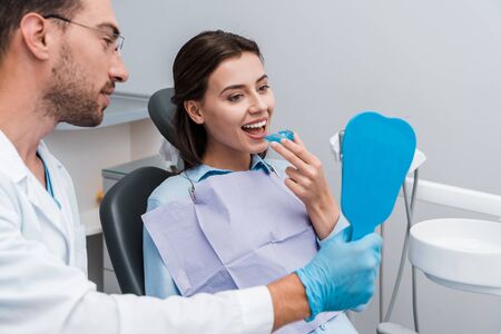selective focus of dentist in glasses holding mirror near attractive woman with retainerの写真素材