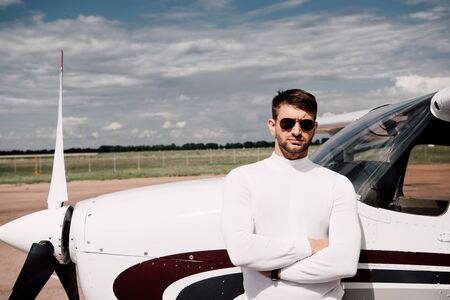 front view of man in sunglasses standing with crossed arms near plane in sunny day