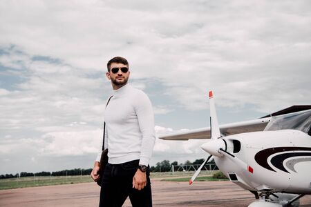 man in sunglasses with bag standing near plane under cloudy sky