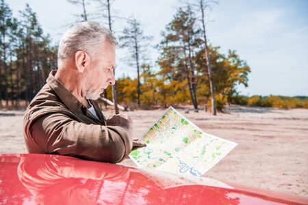 focused senior tourist standing near red car and looking at map in sunny dayの写真素材