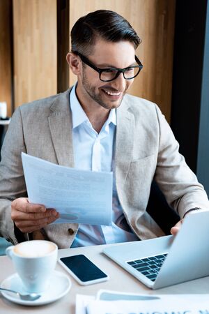 smiling businessman in glasses holding document and typing on laptop keyboard in cafeの写真素材