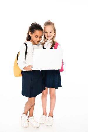 full length view of two multicultural schoolgirls using laptop isolated on whiteの写真素材