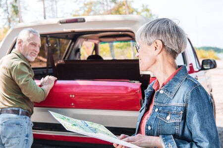 senior man standing near red car while wife holding map in sunny day
