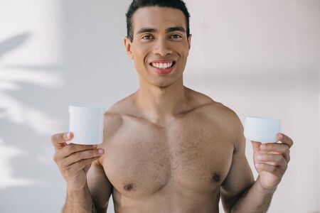 shirtless young man holding two containers for cosmetic cream, smiling and looking at camera