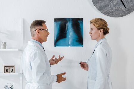 doctor in glasses gesturing near x-ray while looking at attractive coworkerの写真素材