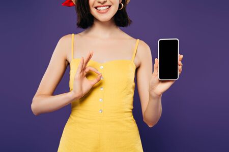 cropped view of smiling girl in holding smartphone with blank screen and showing ok sign isolated on...