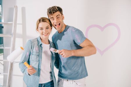 happy young couple looking at camera while holding pink paintbrush and paint rollerの写真素材