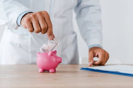 cropped view of doctor in white coat putting dollar banknote in piggy bank の写真素材