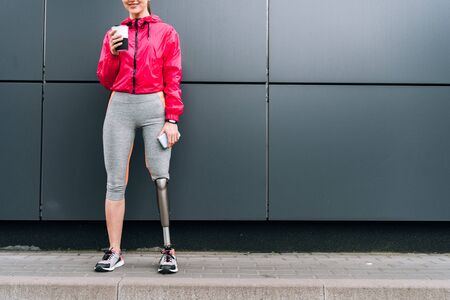 cropped view of smiling disabled sportswoman holding cup and smartphone on streetの写真素材