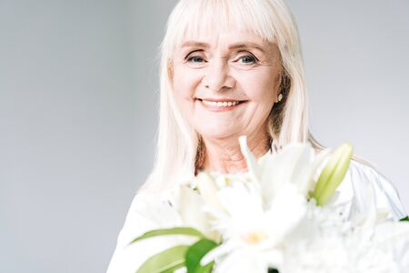 happy blonde senior woman in white clothes holding flowers isolated on grey