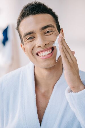 handsome young man in bathrobe applying shaving cream on face while smiling and looking at cameraの写真素材
