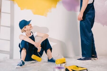 attractive young painter in uniform sitting on floor and looking at man in uniformの写真素材