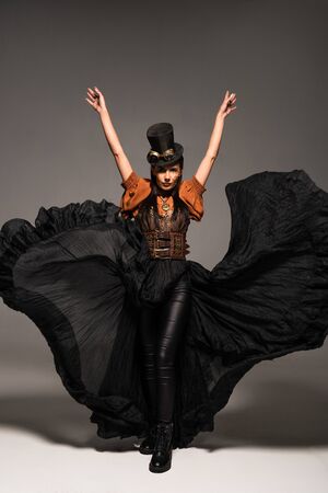 full length view of attractive steampunk woman in top hat with goggles standing with hands up on greyの写真素材