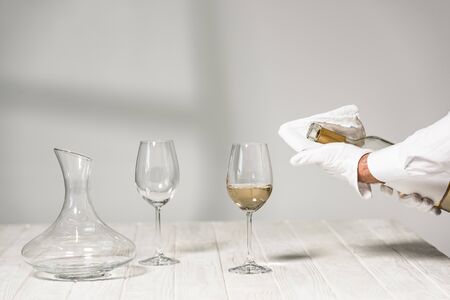 cropped view of water in white glove holding bottle of wine near wine glasses on wooden table