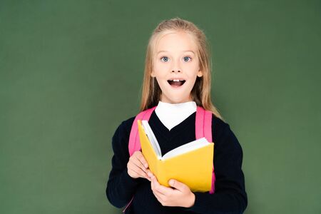 surprised schoolgirl holding book and looking at camera while standing near green chalkboardの写真素材