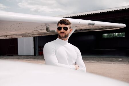 pensive bearded man in sunglasses standing with crossed arms near plane
