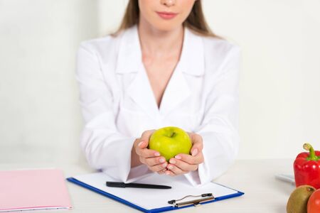 partial view of dietitian in white coat holding apple at workplaceの写真素材