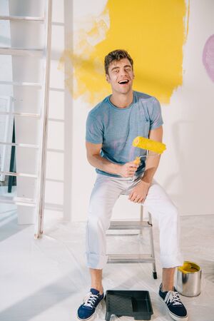 handsome young man sitting on ladder, holding yellow paint roller and smiling at cameraの写真素材