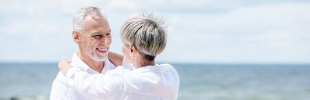 panoramic view of smiling senior couple embracing near river under blue sky