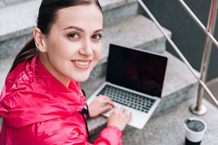 smiling sportswoman using laptop with blank screen on stairs on street