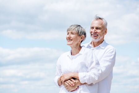 happy senior couple in white shirts embracing under blue skyの写真素材