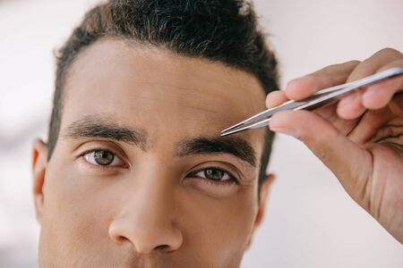 close up view of handsome man plucking eyebrows with tweezer and looking at camera
