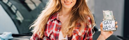 panoramic shot of happy girl and standing with hand on hip and holding jar with car wash letters and moneyの写真素材